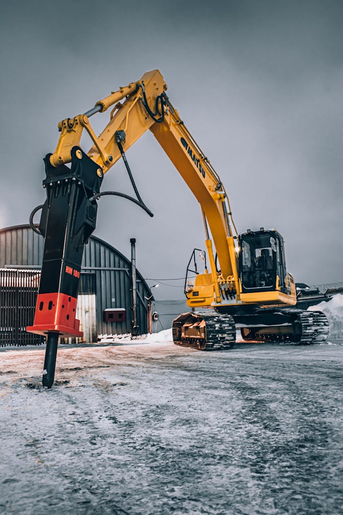 A yellow excavator with a hydraulic breaker parked on a snowy construction site.